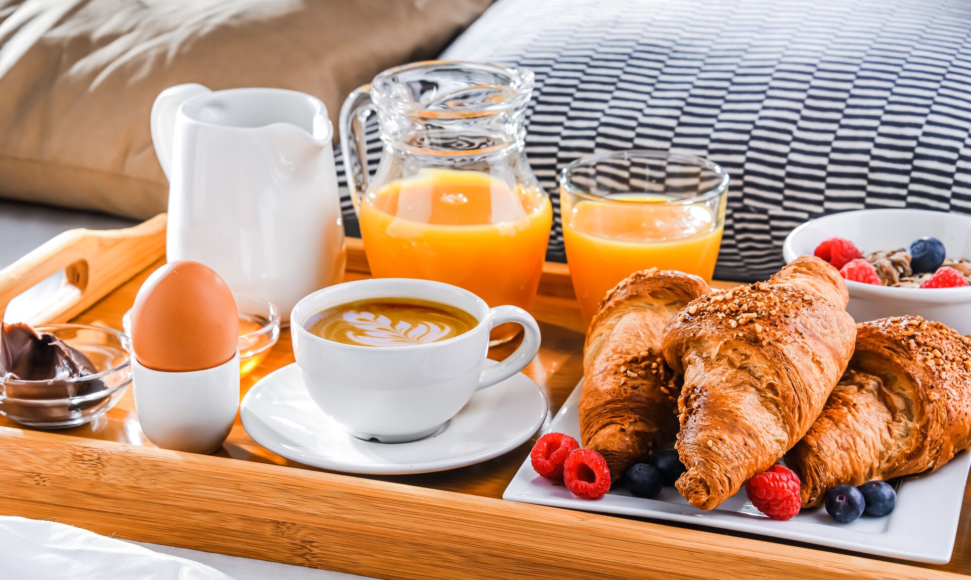 Ein Frühstückstablett mit Croissants, Kaffee, Orangensaft und einem gekochten Ei auf einem Bett.